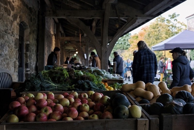 Apples, squash and leafy greens in crates on a market stall under a timber roof, autumn shoppers blurred behind
