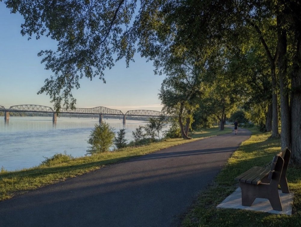 A paved riverside trail curving under tall trees past a wooden bench, a steel truss bridge over calm water in morning light