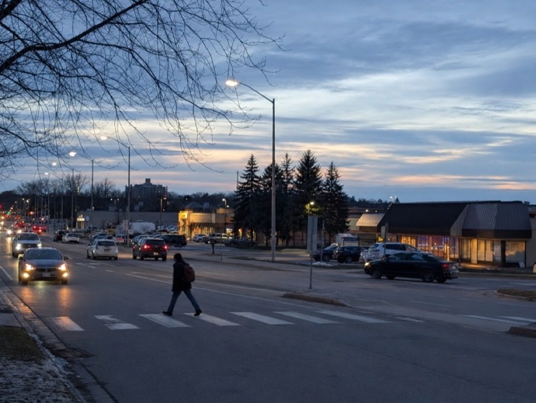 A wide suburban road at dusk lined with single-storey plazas, car headlights, bare trees and a person with a backpack at a crosswalk